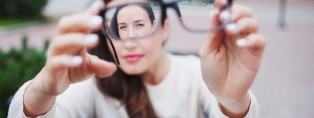 Closeup portrait of young women with glasses. She has eyesight problems and is squinting his eyes a little bit. Beautiful girl is holding eyeglasses right in front of camera with two hand